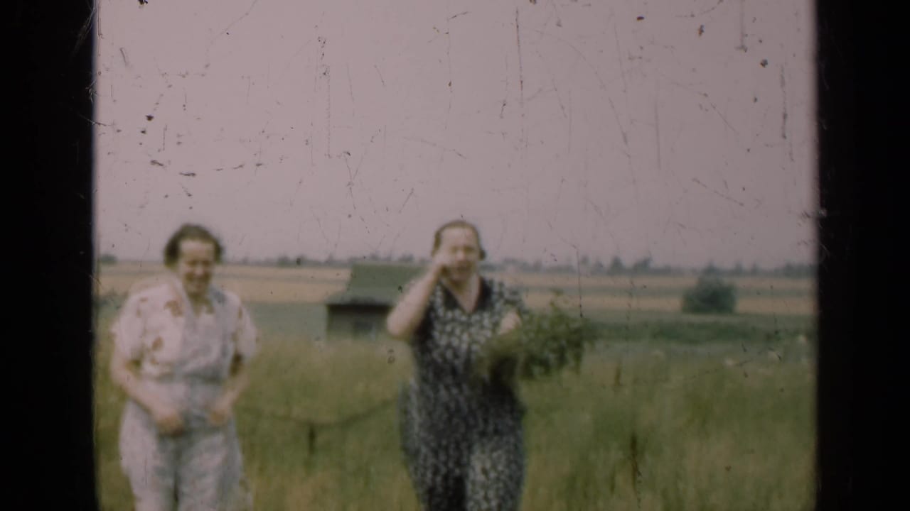 Two women stand in a grassy field with distant farmland behind them.
