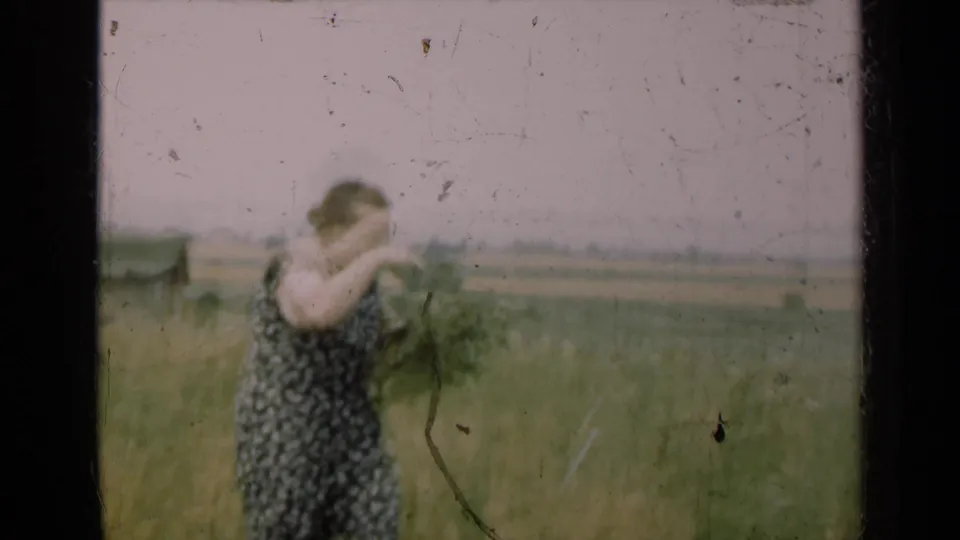 A woman in a dark patterned dress turns with gathered plants in a rural Illinois field, captured in soft 8mm home-movie focus.
