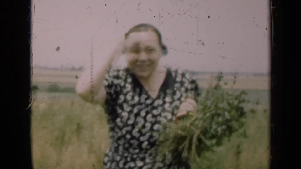 A woman in a patterned dress holds a bundle of green plants and smiles toward the camera in a rural Illinois field.