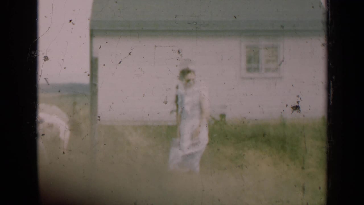 Woman in a pale dress walking across a grassy yard near a light farmhouse.