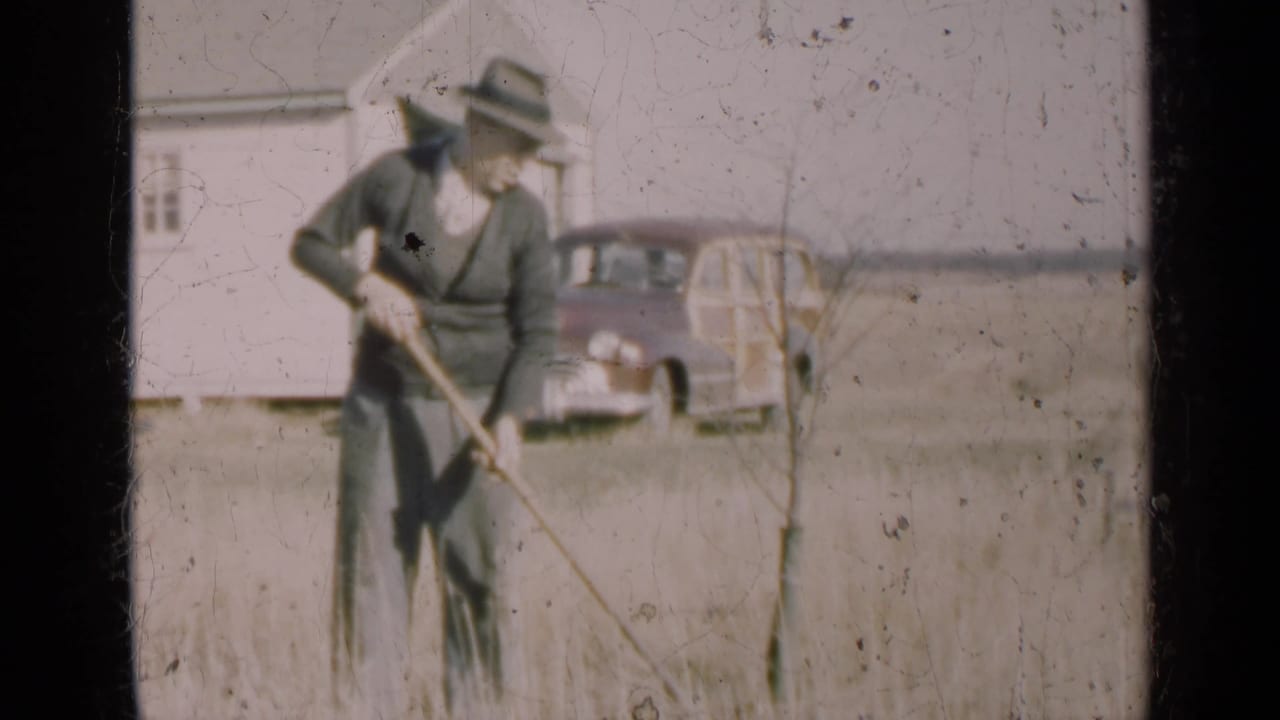 Man in a fedora using a hoe in dry grass with a farmhouse and car behind him.