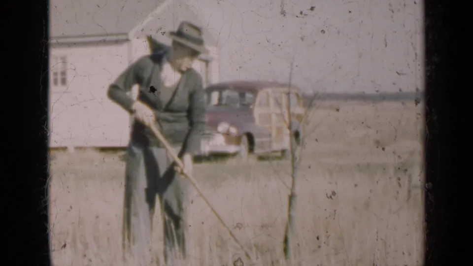 A man in a dark outfit and fedora works dry ground with a hoe outside a farmhouse in 1947 Illinois.