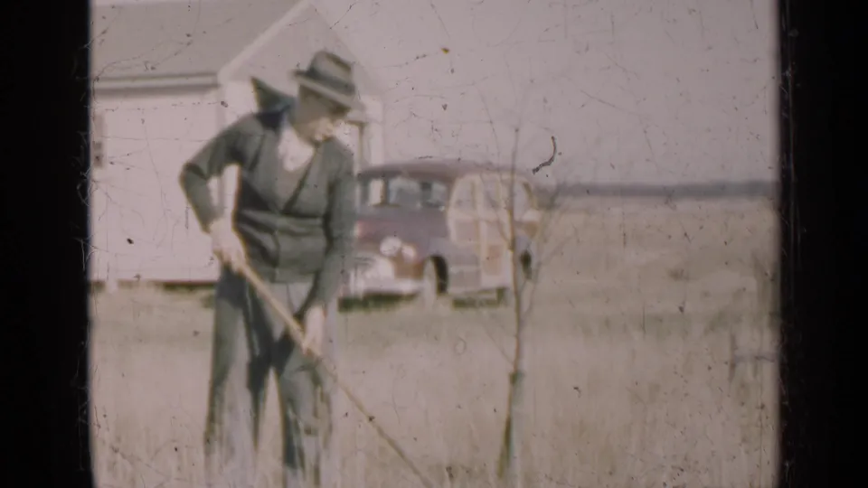 A 1947 Illinois yard scene shows a man pausing with a hoe near a farmhouse and parked older car.