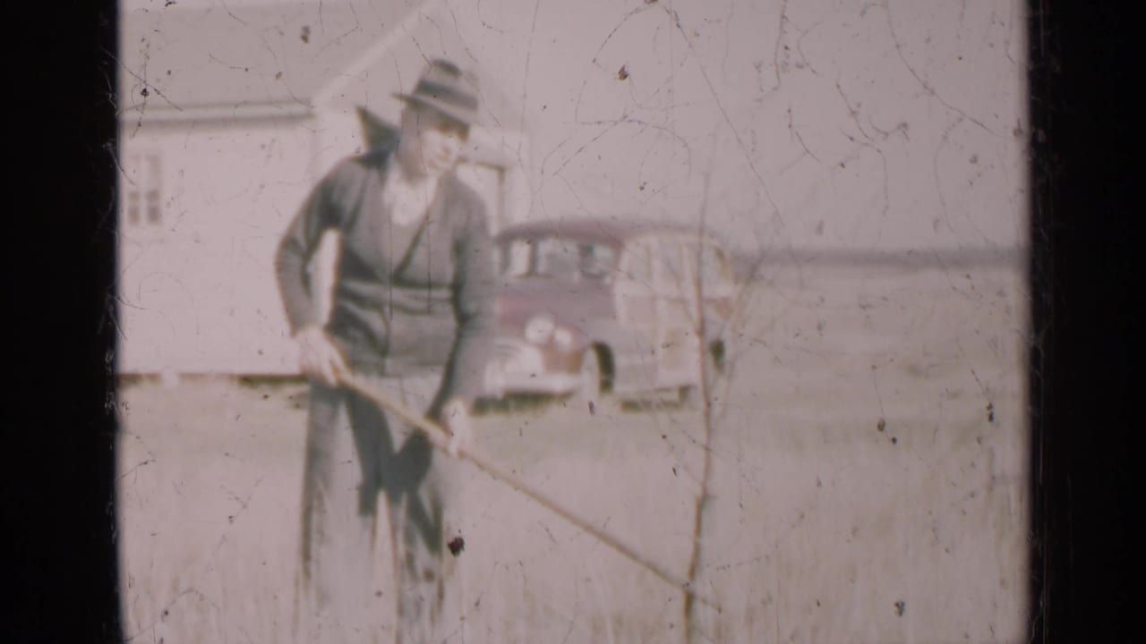 Man in a hat raking tall grass near a white house and parked vintage car.