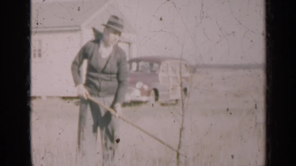 1947 Illinois home-movie frame showing a man in a fedora raking tall grass with a white house and station wagon behind him.