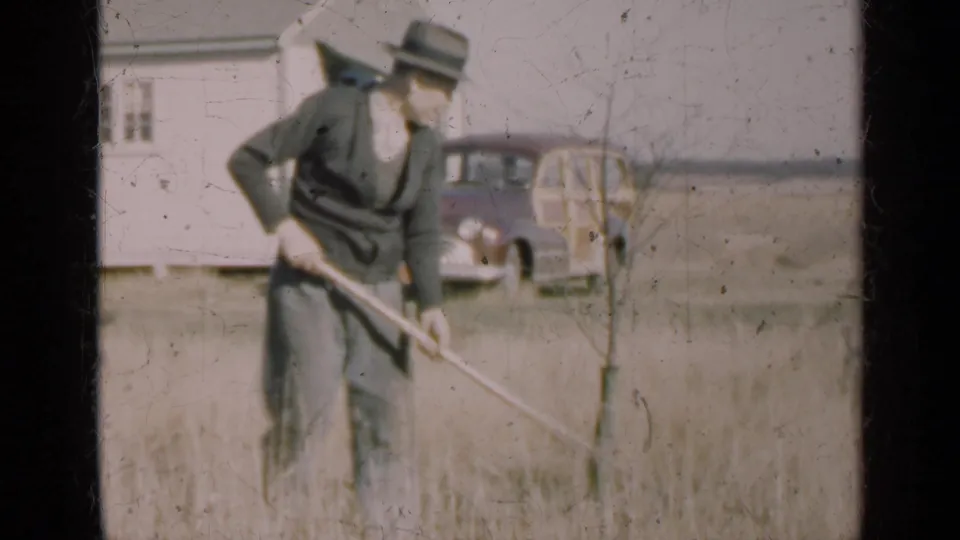 A man in 1947 Illinois appears to rake grass near a small tree, with a white house and vintage station wagon in the background.