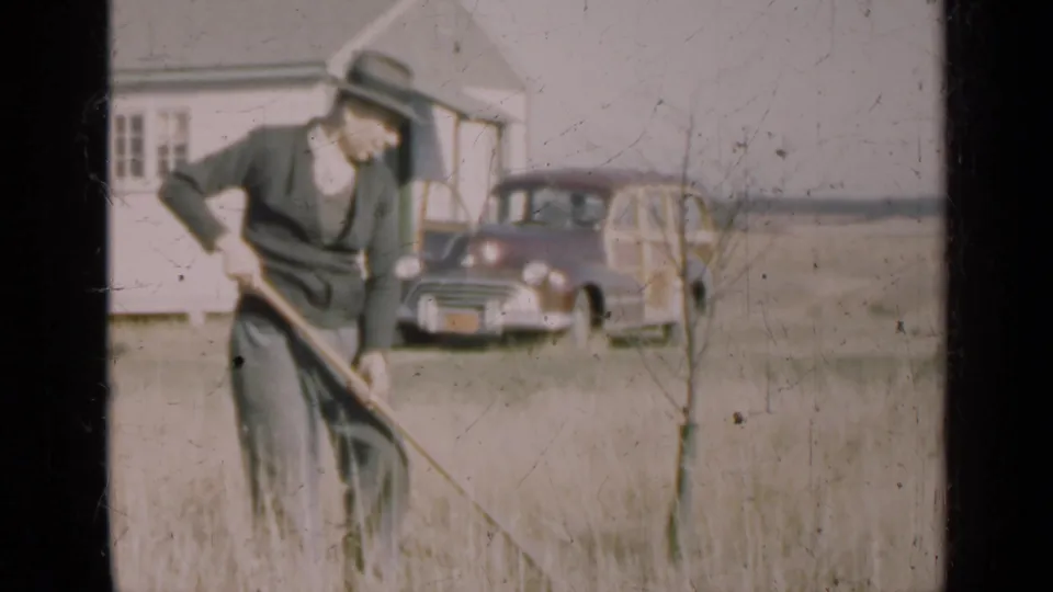 Illinois 1947 home-movie still of a man leaning over a rake in tall grass, with a modest house and station wagon behind him.