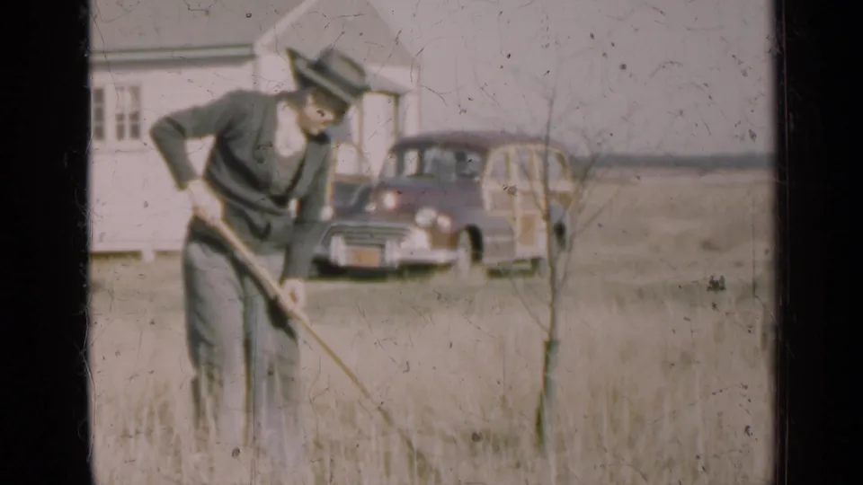 A 1947 Illinois 8mm frame shows the man continuing to rake tall grass near a small tree, house, and vintage station wagon.