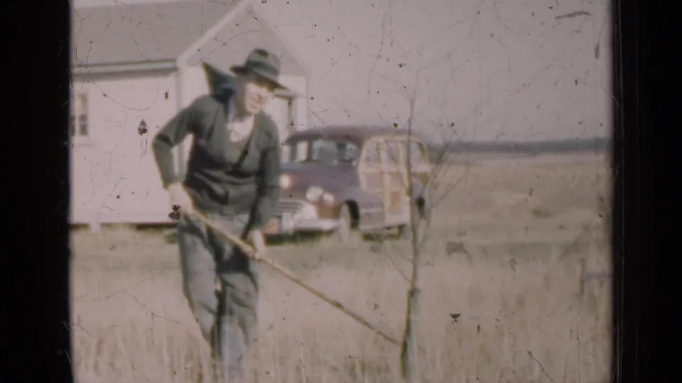 Soft 8mm frame of a man in dark clothing tending a small tree in an Illinois yard, backed by a house and mid-century wagon.