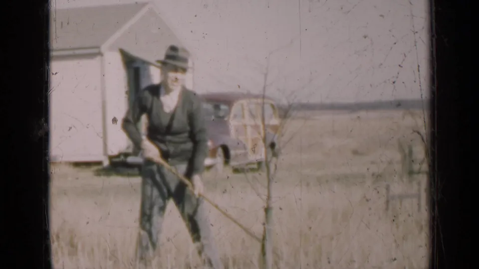 A 1947 Illinois home-movie frame shows the man finishing a motion around the sapling, framed by tall grass, house, and wagon.