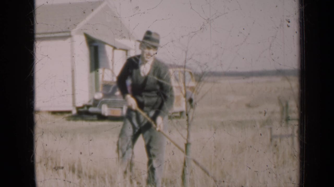 Man in a fedora works with a long-handled tool in a dry field near a white house and parked car.
