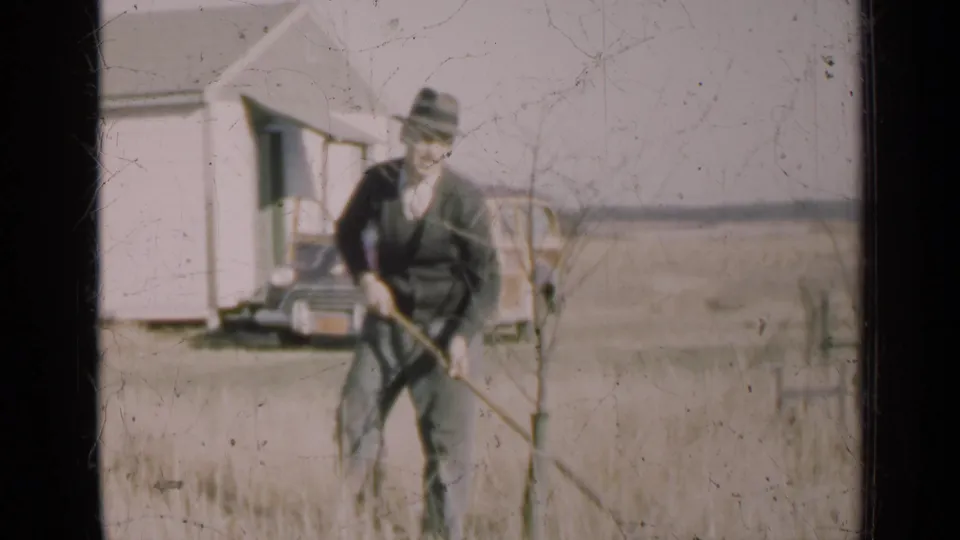 In rural Illinois home-movie footage from 1947, a man in a fedora tends ground near a sapling, with a white house and 1940s car behind him.