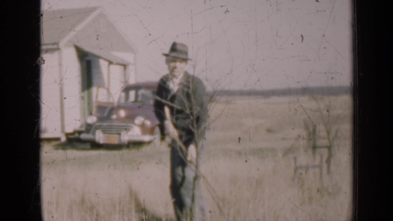 Man stands in a field holding a long-handled tool, with a vintage car and small white house behind him.