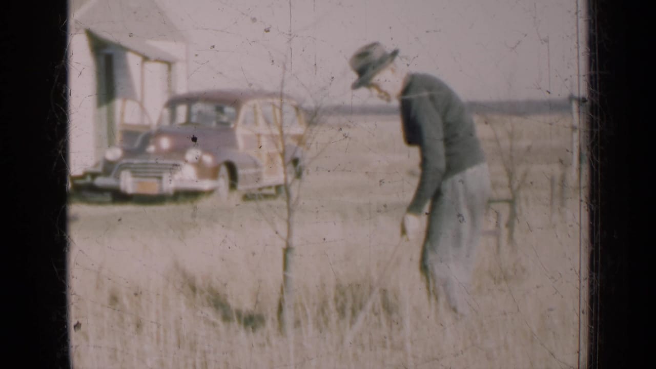 In 1947 Illinois home-movie footage, a man works with a rake in a dry field while a wood-paneled station wagon sits near a simple building.
