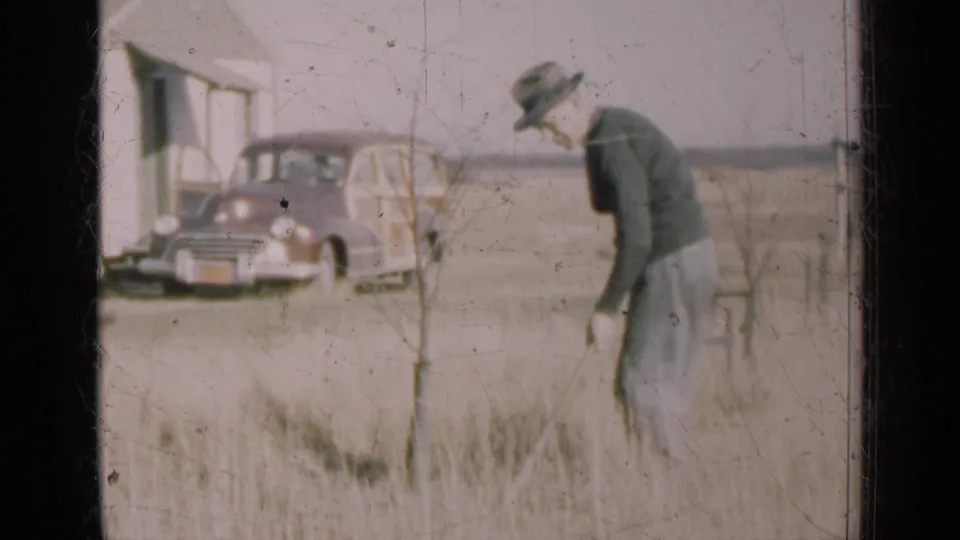 In 1947 Illinois home-movie footage, a man works with a rake in a dry field while a wood-paneled station wagon sits near a simple building.