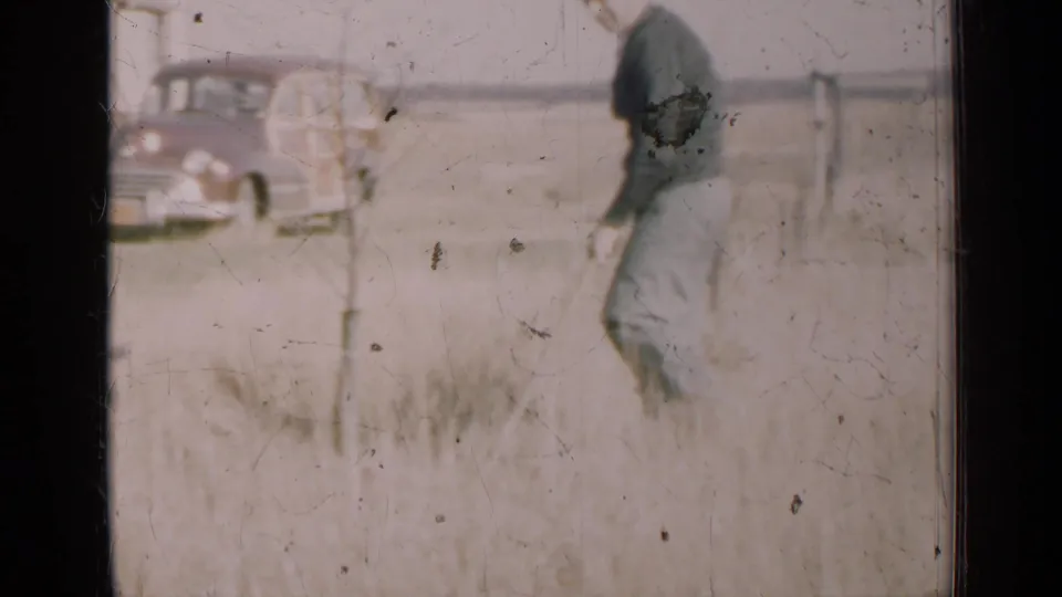 The home-movie frame shows the man closer to camera as he moves through the field, with the vintage station wagon partly seen at left.