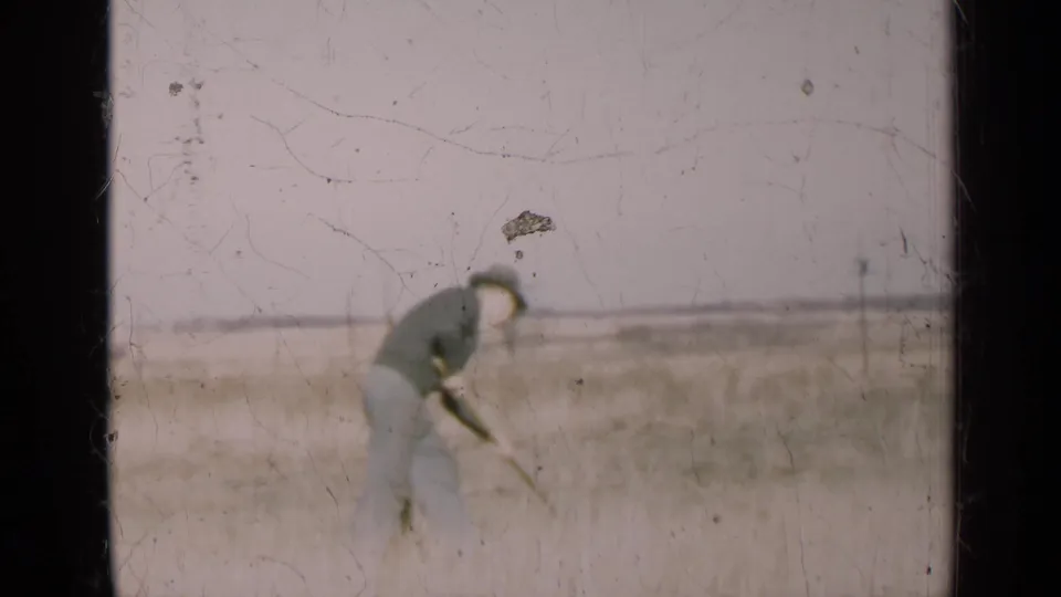 A distant view shows the man bent over his rake in an open Illinois field, framed by soft 8mm grain, scratches, and pale sky.