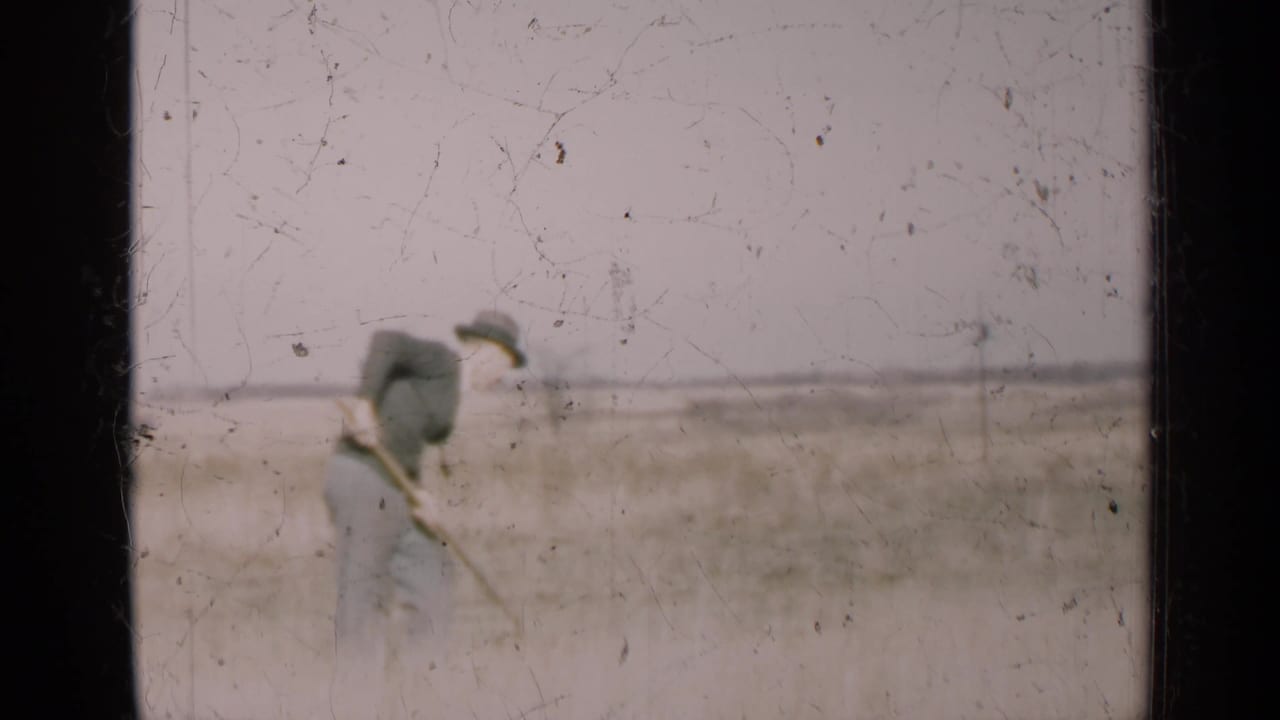 Man in a dark shirt and hat holds a scythe in a dry open field.