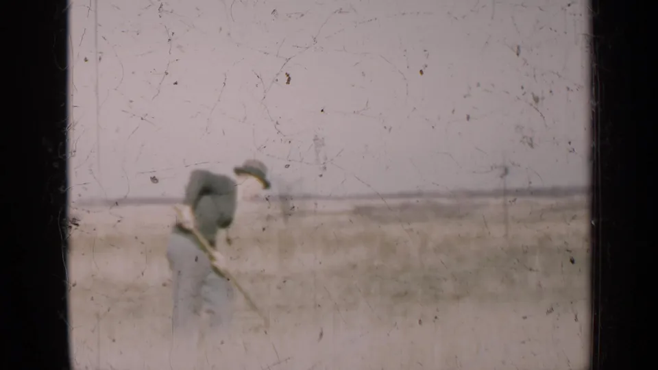 In 1947 Illinois home-movie footage, a man stands with a scythe in a dry field under a pale sky.