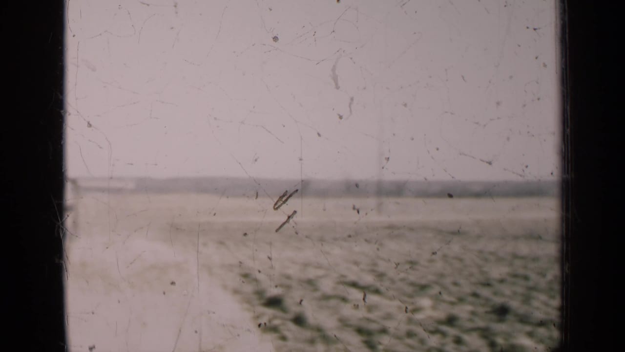 Open field with a faint track and distant poles behind dark window dividers.