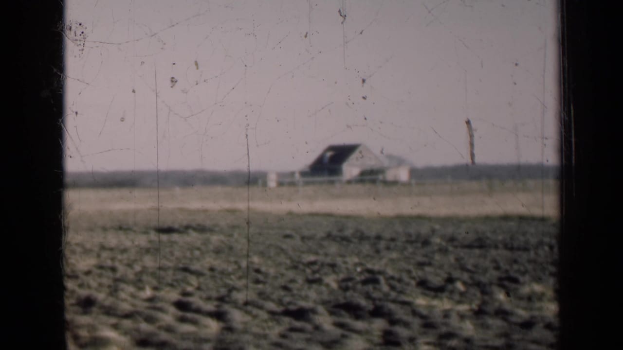 Distant farm building beyond an open field, framed by dark 8mm film borders.