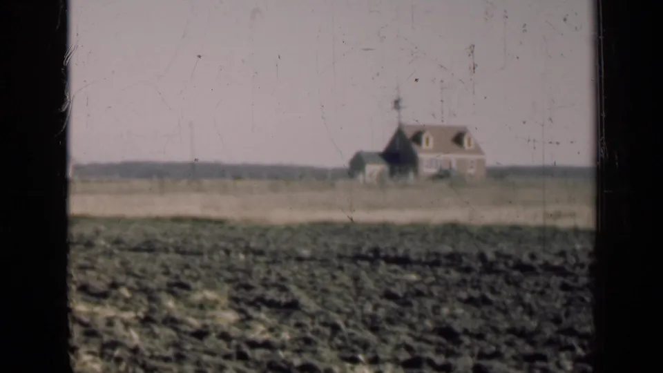 1947 Illinois 8mm frame showing a distant rural farmhouse beyond a field, partly framed by a dark window edge.