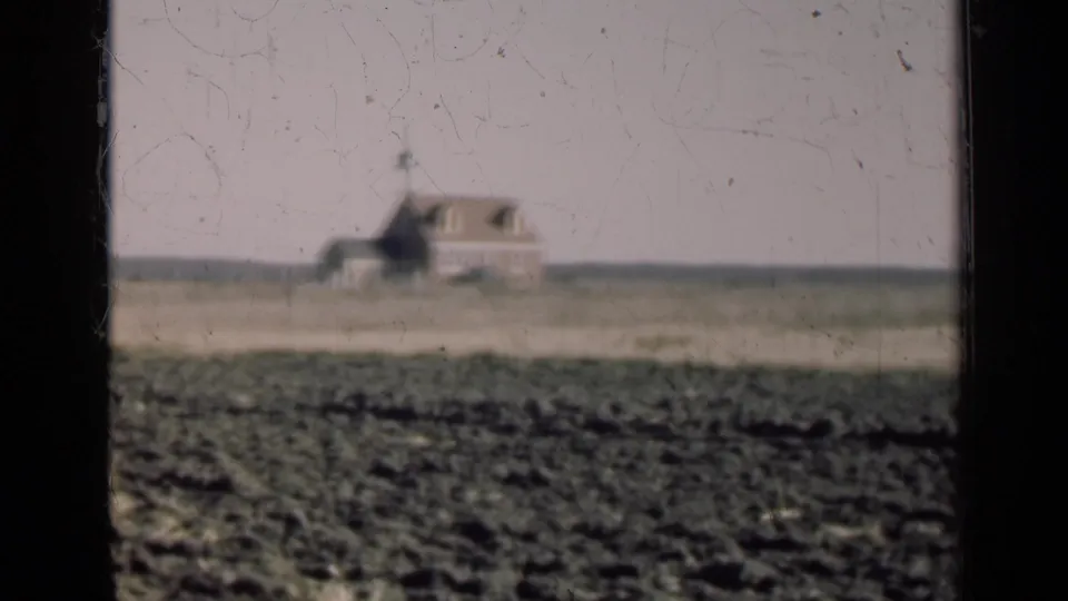 Soft 8mm view of a rural Illinois farmhouse across open fields, with a dark window divider cutting through the frame.