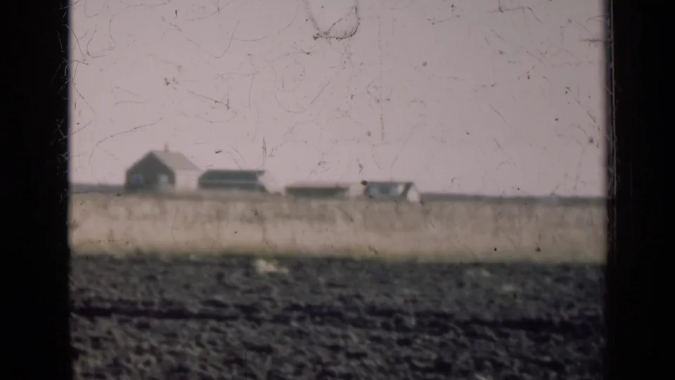 Distant Illinois farm buildings and possible equipment appear across a plowed field under a pale sky.