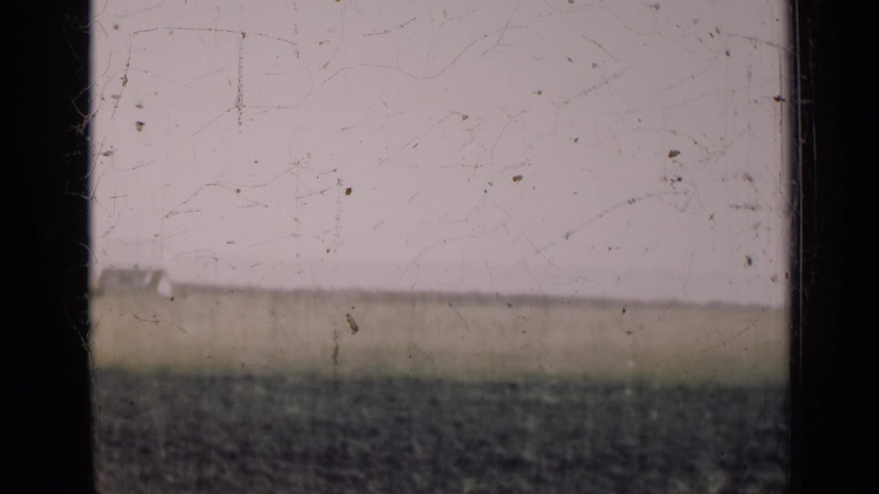 A flat Illinois field and low horizon sit behind a central dark frame bar, with visible scratches and grain from the 8mm source.