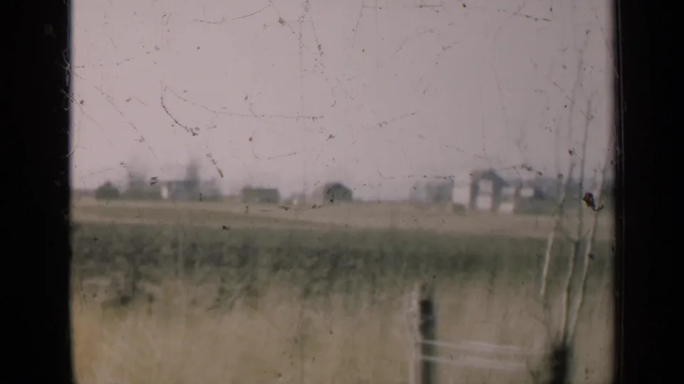 A rural Illinois field shows grasses, a fence post, and distant farm buildings, viewed through a dark frame with visible aged-film marks.