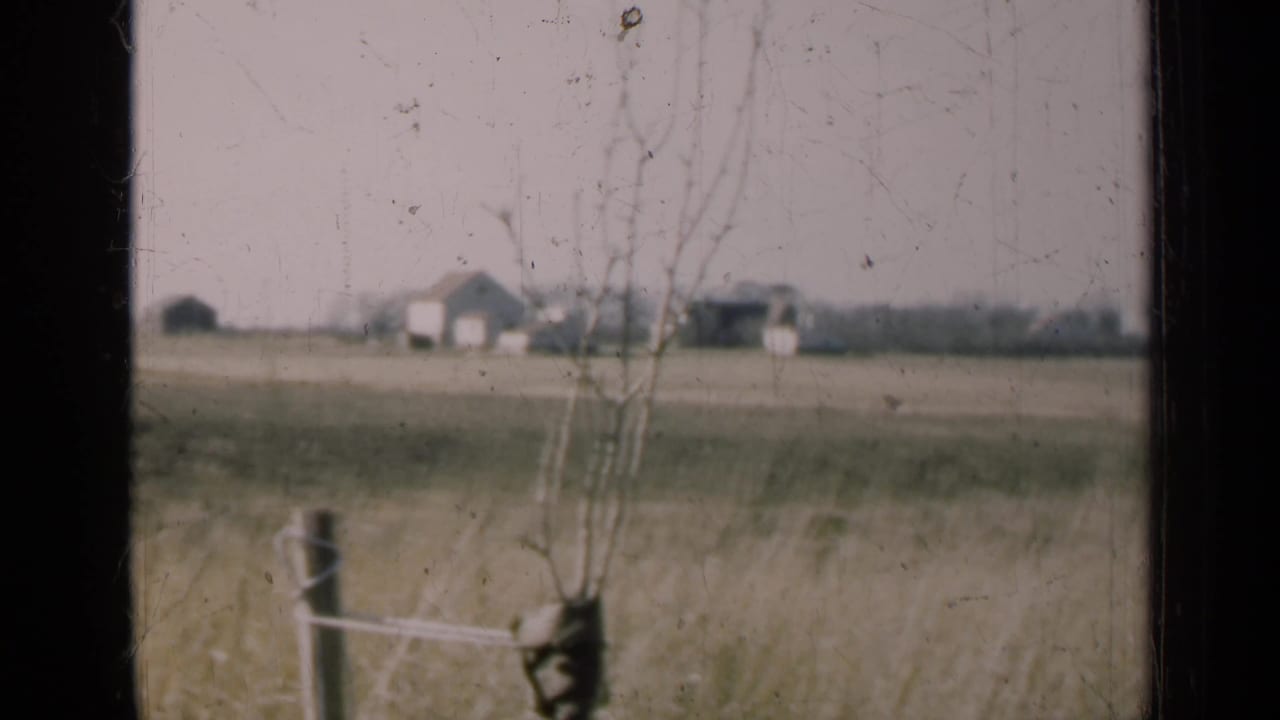 A dusty 8mm view of an Illinois farmstead across open fields, with bare trees and a fence in the foreground.
