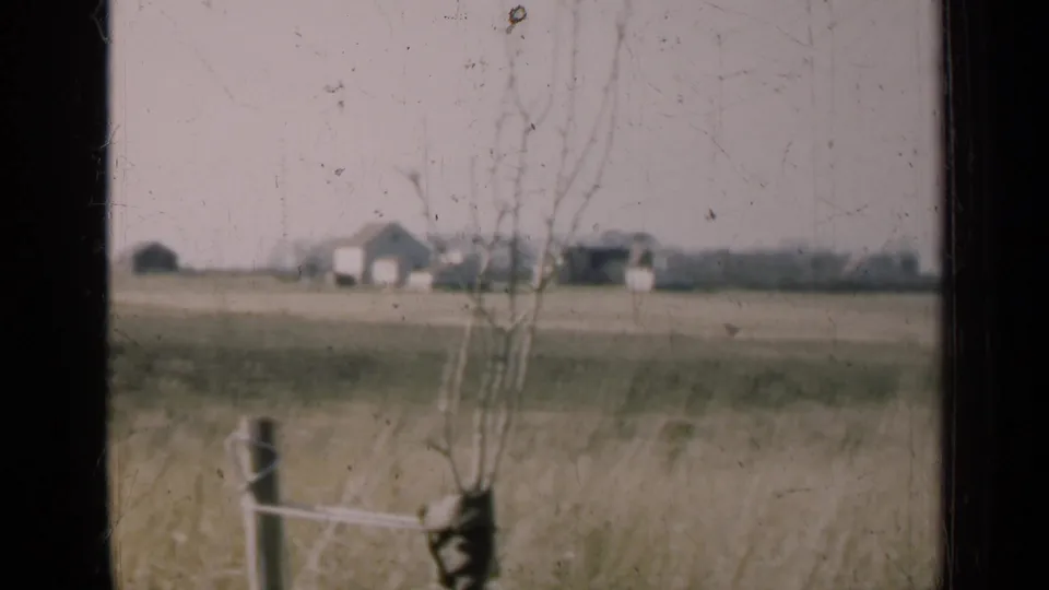 A dusty 8mm view of an Illinois farmstead across open fields, with bare trees and a fence in the foreground.
