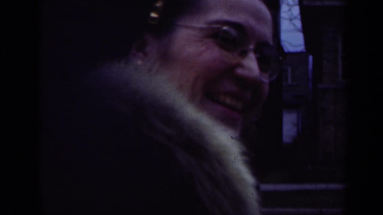 1947 Illinois home-movie frame of a smiling woman in glasses and a fur-collared coat near a residential street or entry.