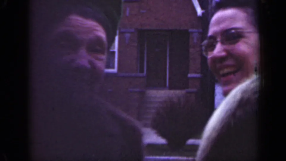 1947 Illinois home-movie frame of two smiling women, one in glasses and a light fur-collared coat, outside brick homes.