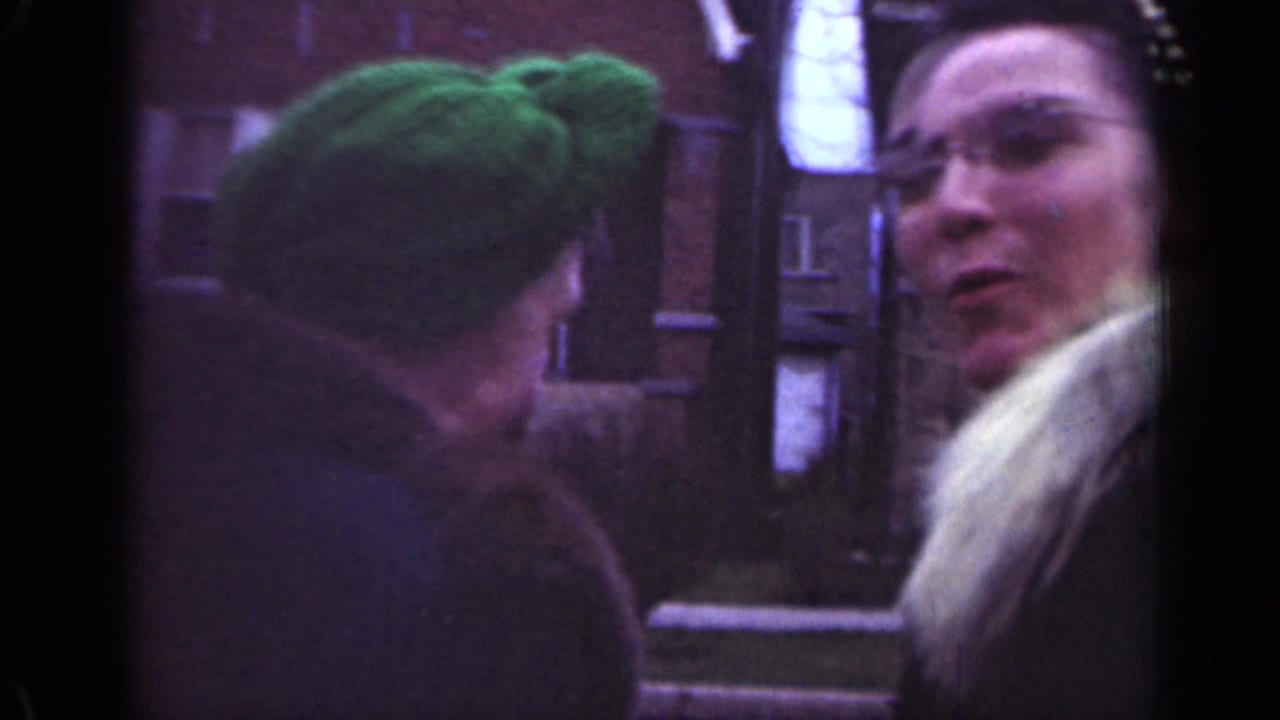 Two adult women stand on a street near brick buildings, one in a green hat and the other in a fur-trimmed coat.