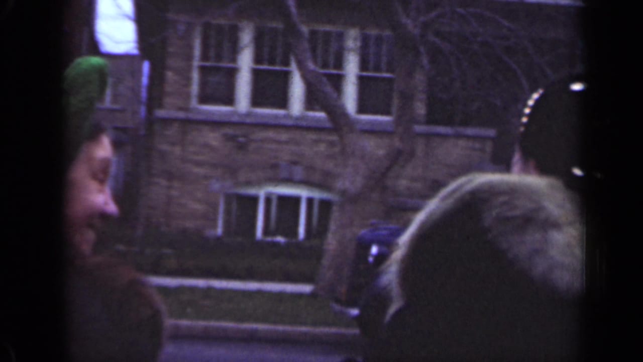In the Illinois street scene, the women turn beside a parked car, with brick buildings, bare branches, and aged 8mm grain visible.
