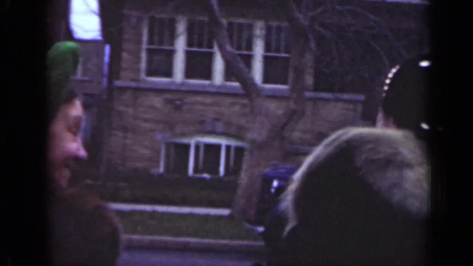 In the Illinois street scene, the women turn beside a parked car, with brick buildings, bare branches, and aged 8mm grain visible.