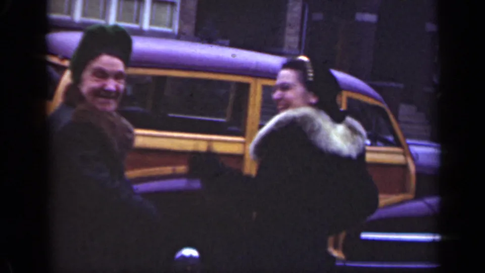 In 1947 Illinois, two women laugh beside a wood-panel station wagon, with one woman facing the camera and the other in a fur-trimmed coat.