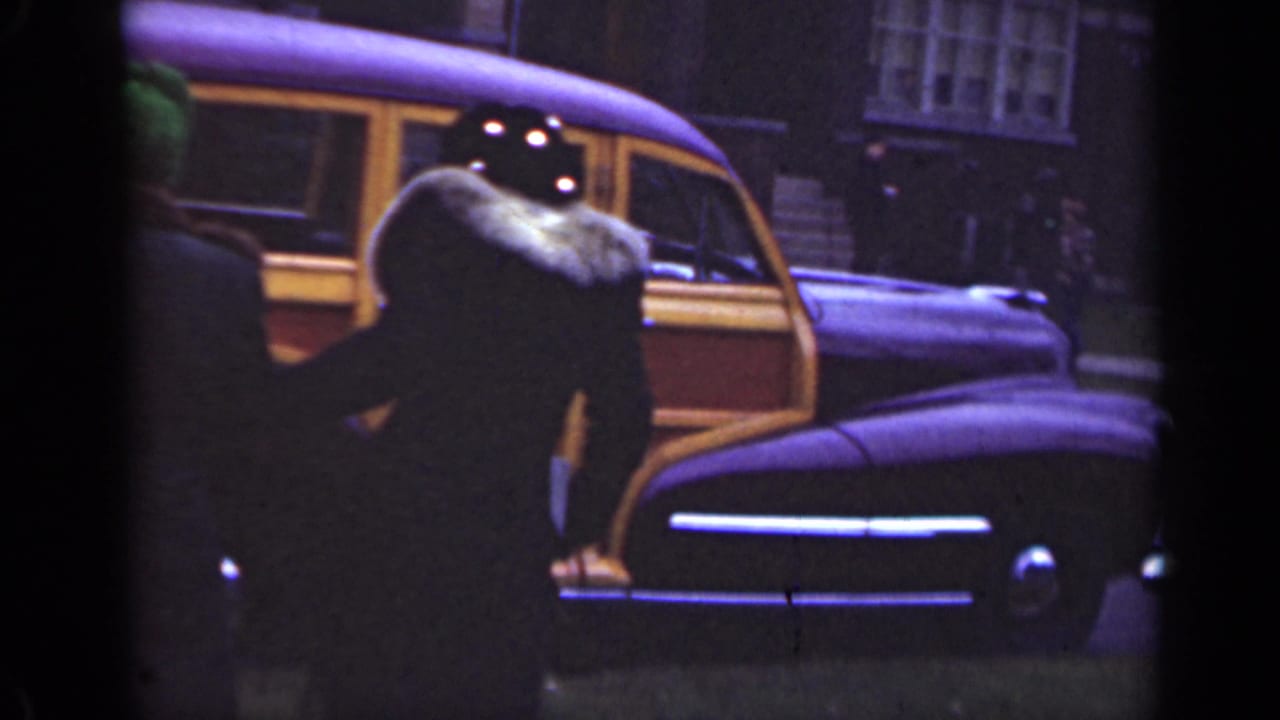 Two adults near a wood-paneled station wagon on a dim street.