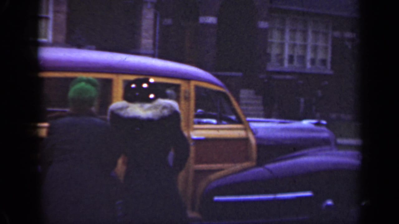 The 1947 Illinois home-movie frame shows an adult in a fur-collared dark coat beside a wood-paneled station wagon as another person stands nearby.