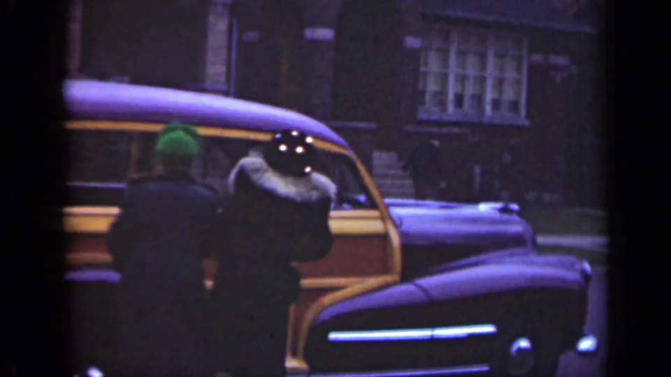 Two adults are seen from behind beside a wood-paneled station wagon in the Illinois 1947 street scene.