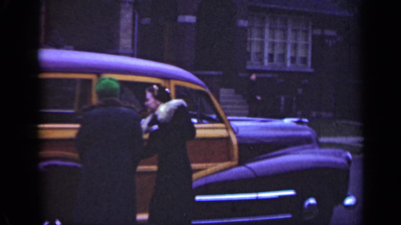 Woman and another person stand beside a wood-panel station wagon on a street.