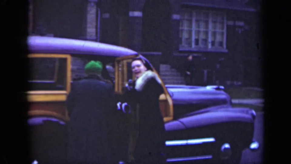 In 1947 Illinois footage, the woman turns and smiles toward the camera beside the wood-panel station wagon, with a brick building behind her.