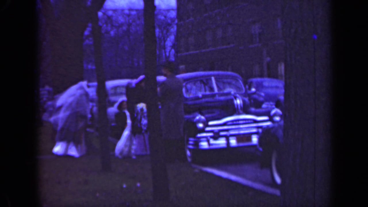 Bride and adults stand near a parked mid-century car on a tree-lined street.