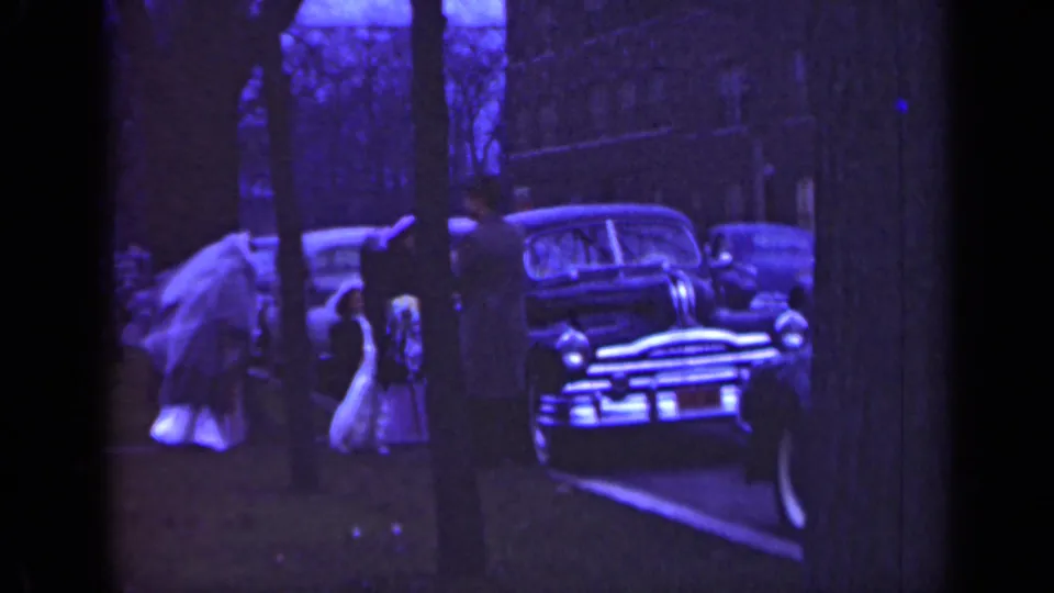 1947 Illinois home-movie frame showing a bride in white and adults near a parked mid-century car outdoors.
