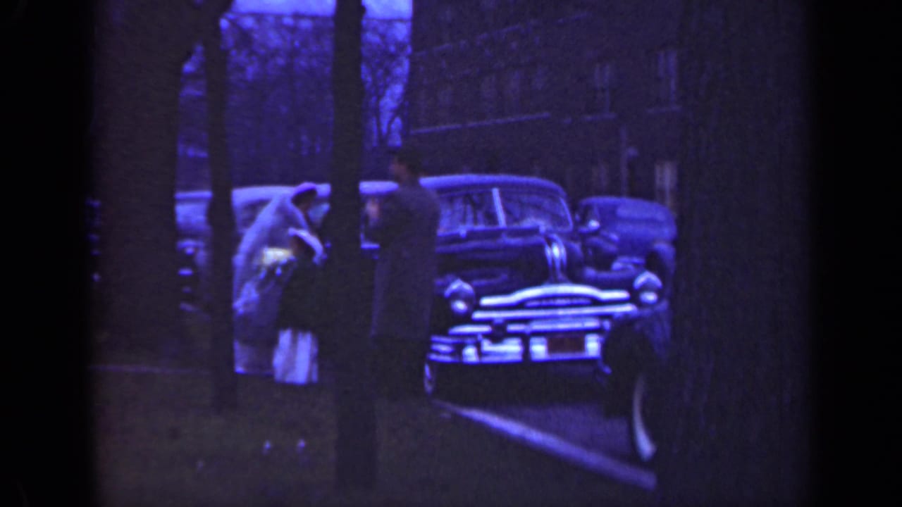 Adults and a bride stand partly behind trees beside a parked car.