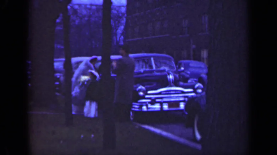 Wedding-party figures in 1947 Illinois appear near a parked car, partially obscured by trees and soft 8mm focus.