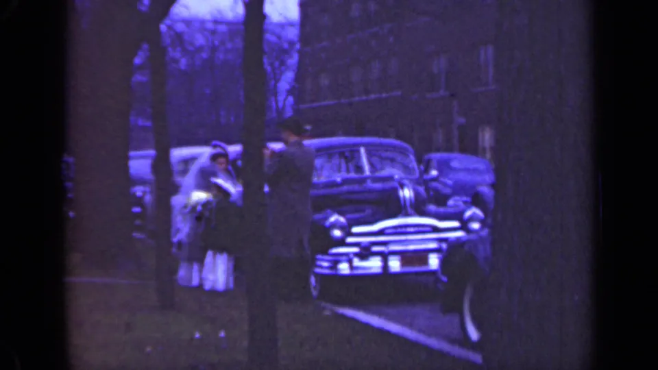 Home-movie frame from Illinois in 1947 showing a bride and adults grouped near a mid-century car at the curb.