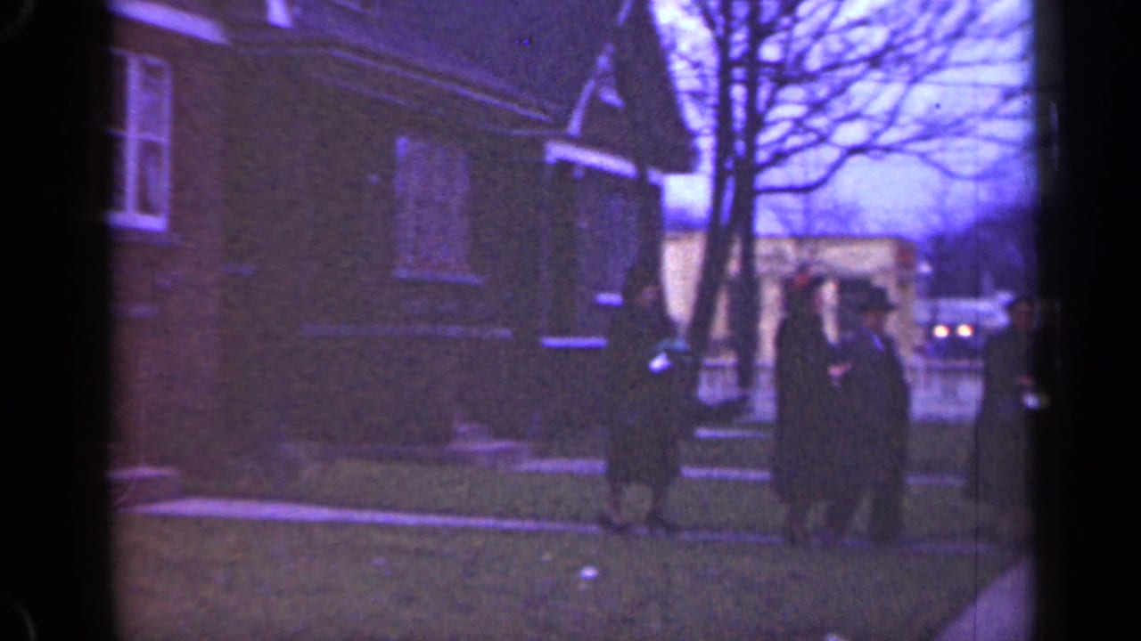 Dark-coated people walk near a brick house and sidewalk.
