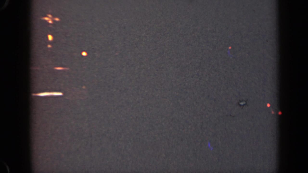 Dark grainy frame with soft orange lights and a small white streak near the left side.